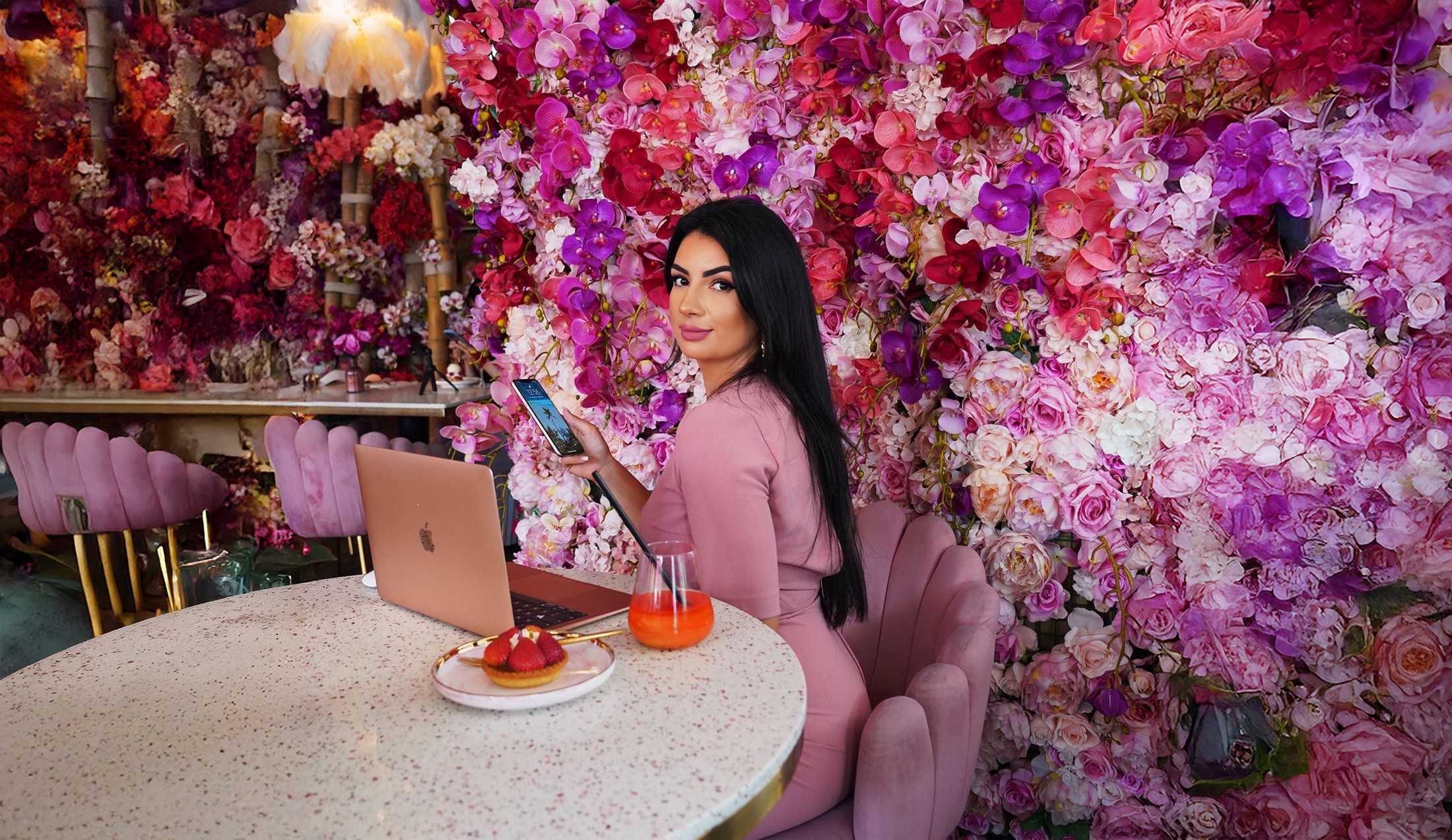 Sobia Mohammed mit Laptop und Smartphone an einem Tisch vor einer pinken Blumenwand